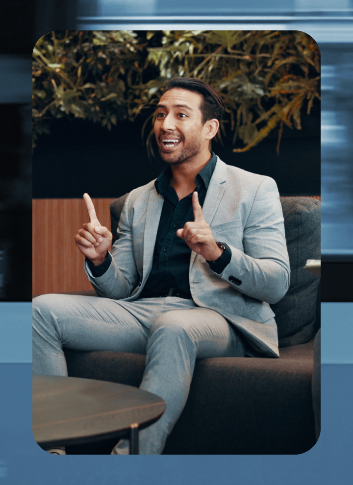 Hombre sentado en un sofá moderno, con traje gris claro y zapatos oscuros, gesticulando con ambas manos en una sala con pared de madera y plantas.