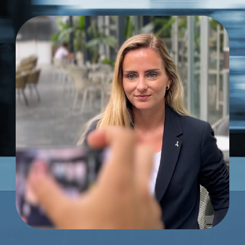 Mujer con chaqueta oscura sentada en una terraza, mientras alguien sostiene un móvil en primer plano.