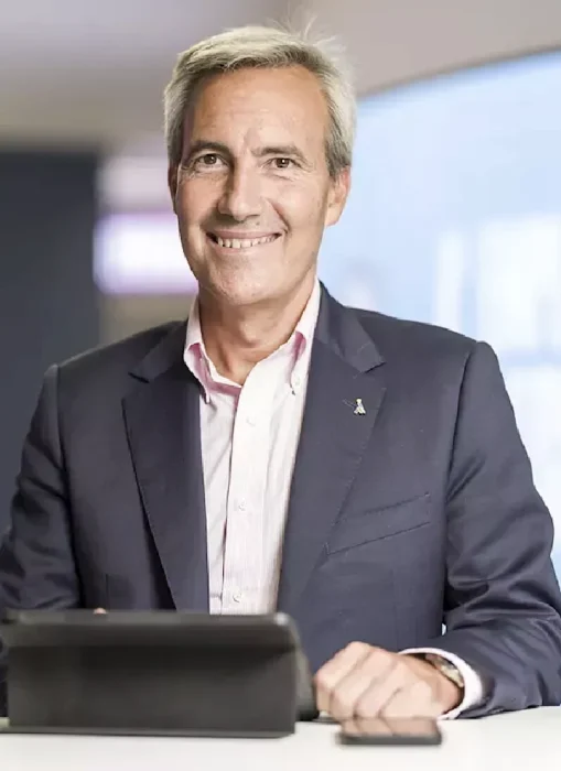 Hombre vestido con chaqueta oscura y camisa clara, sentada frente a una mesa con tablet y teléfono móvil.