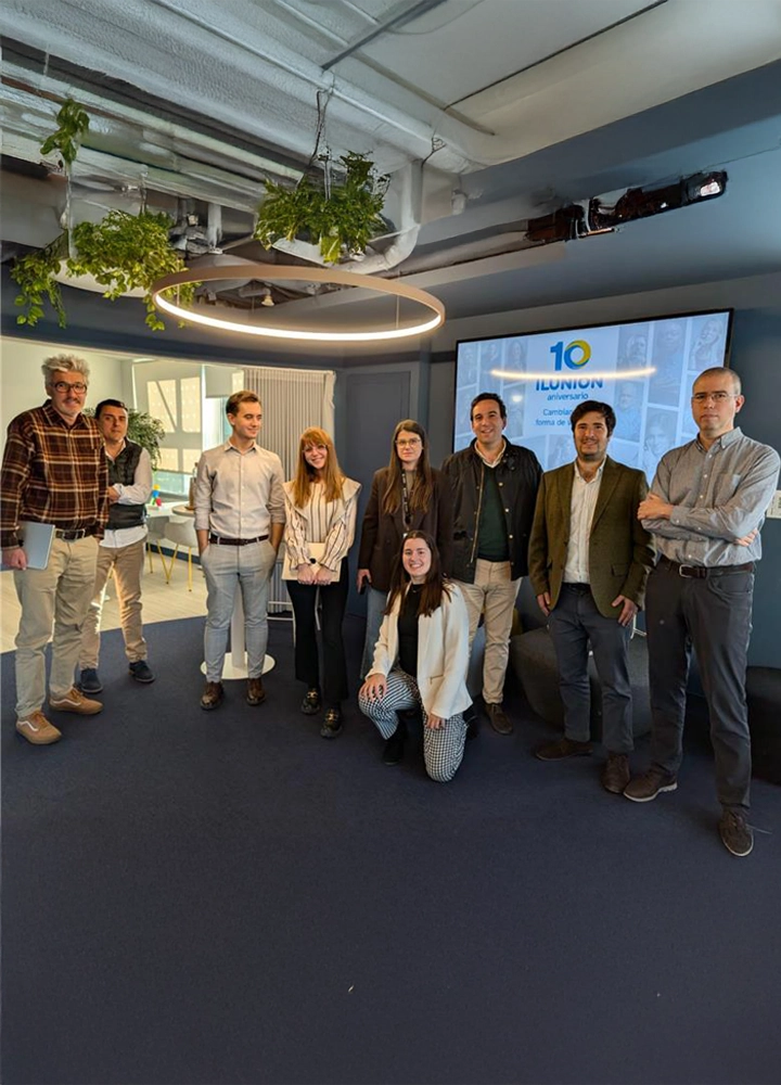 Grupo de personas posando en sala moderna con techo decorado con plantas y lámpara circular. Al fondo, pantalla muestra logo de Ilunion y texto sobre aniversario.