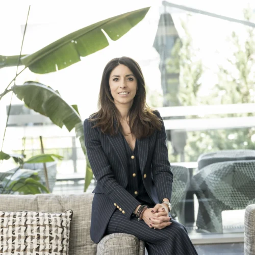 Mujer sentada en un sofá, vestida con traje de chaqueta a rayas oscuras, en un espacio interior luminoso con plantas y mobiliario moderno
