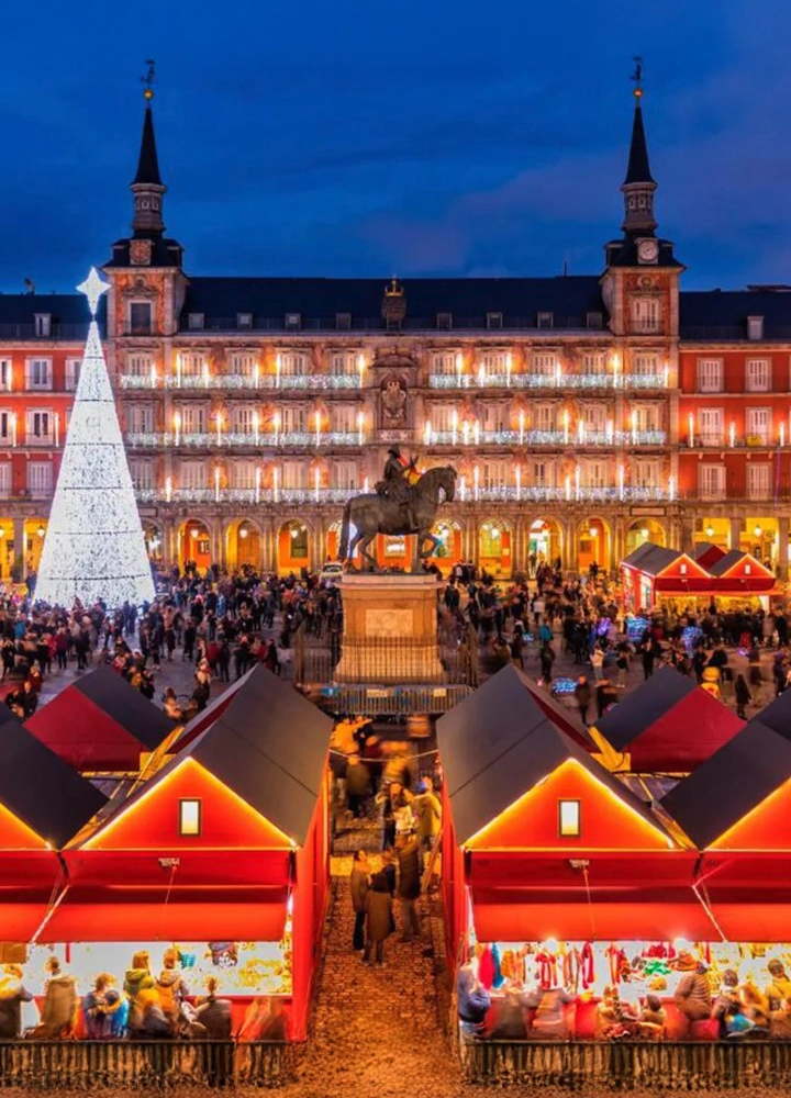 Mercado navideño iluminado en la Plaza Mayor de Madrid, con puestos rojos, un gran árbol de Navidad blanco y edificios históricos al fondo