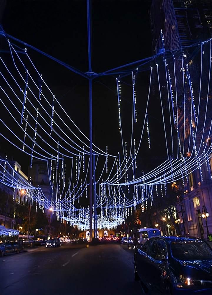 Calle iluminada con decoraciones navideñas azules colgantes, vista nocturna con coches circulando y edificios a ambos lados