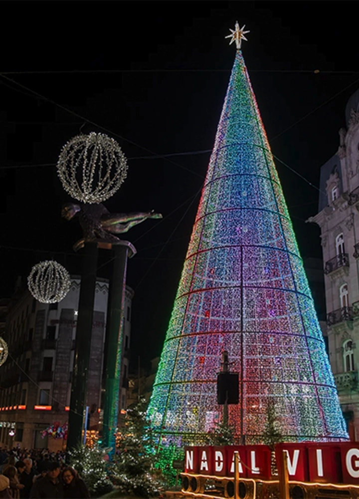 Gran árbol de Navidad iluminado con luces multicolor en una plaza urbana, rodeado de decoraciones esféricas y edificios históricos, con el texto 'Nadal Vigo' en la base