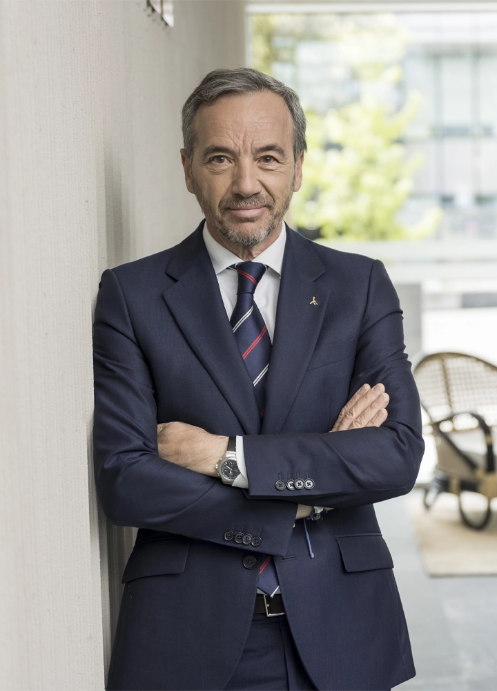 Hombre vestido con traje formal azul marino y corbata a rayas, de pie con brazos cruzados junto a una pared en un espacio moderno con mobiliario