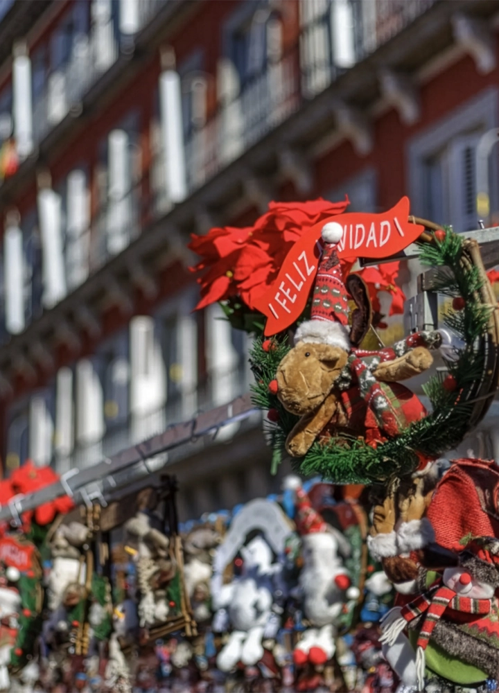 Puesto navideño al aire libre decorado con peluches, coronas y adornos festivos. Destaca un reno con gorro de Papá Noel junto al cartel rojo “¡FELIZ NAVIDAD!”. El entorno urbano con fachada de ladrillo y ventanas blancas refuerza el ambiente alegre y tradicional de las celebraciones navideñas.