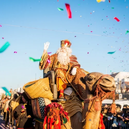 Escena festiva de la Cabalgata de Reyes con Melchor saludando desde un camello adornado con telas y borlas coloridas. El personaje lleva corona, túnica ornamentada y barba blanca, mientras cae confeti sobre la multitud. El cielo despejado y los edificios al fondo enmarcan esta celebración tradicional llena de alegría y simbolismo.
