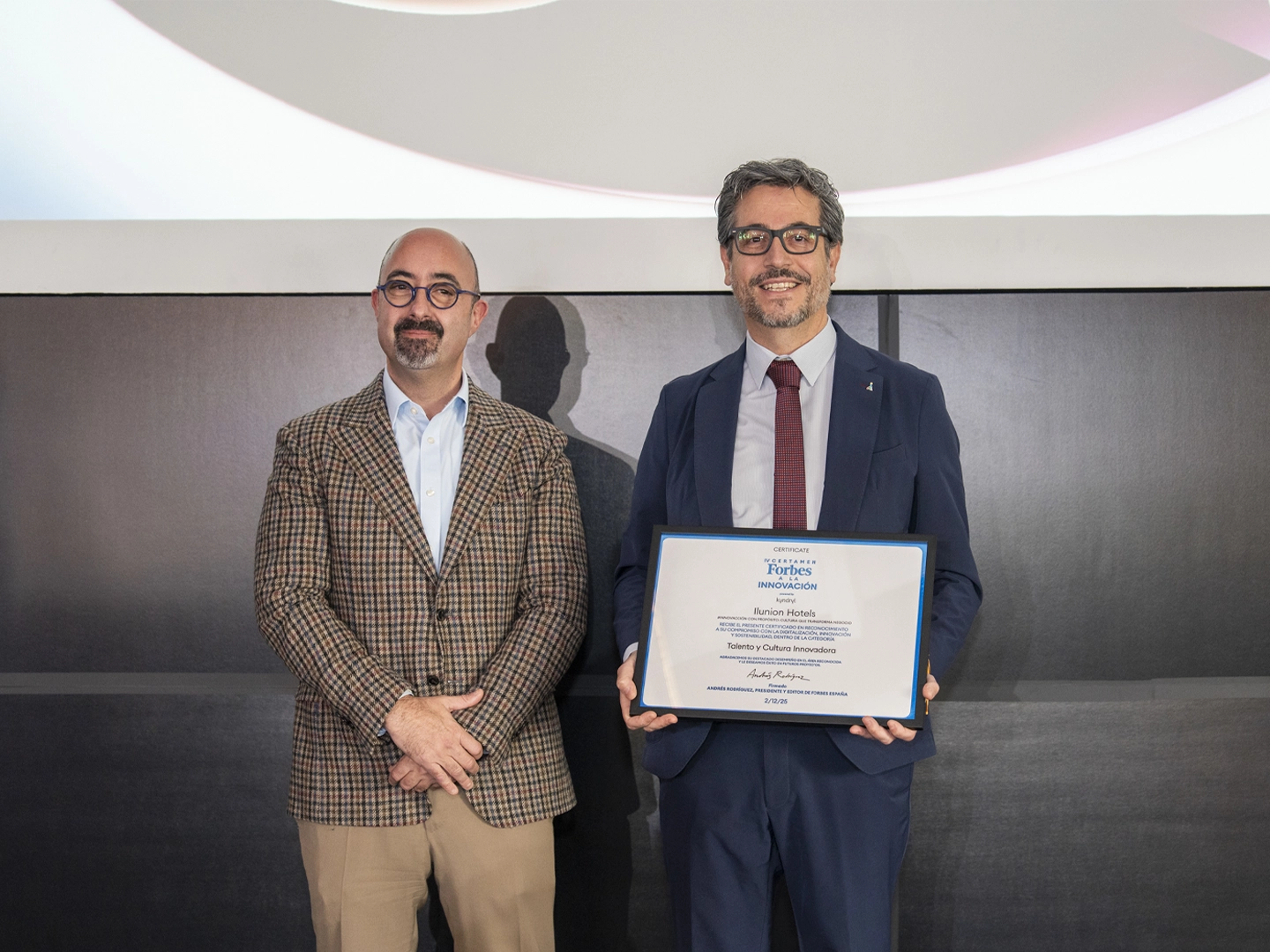 Dos hombres posan en un evento corporativo; una sostiene un diploma enmarcado con el logo de Forbes y texto sobre talento e innovación