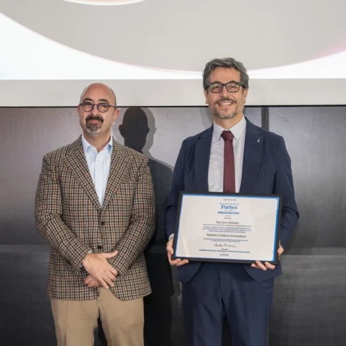 Dos hombres posan en un evento corporativo; una sostiene un diploma enmarcado con el logo de Forbes y texto sobre talento e innovación