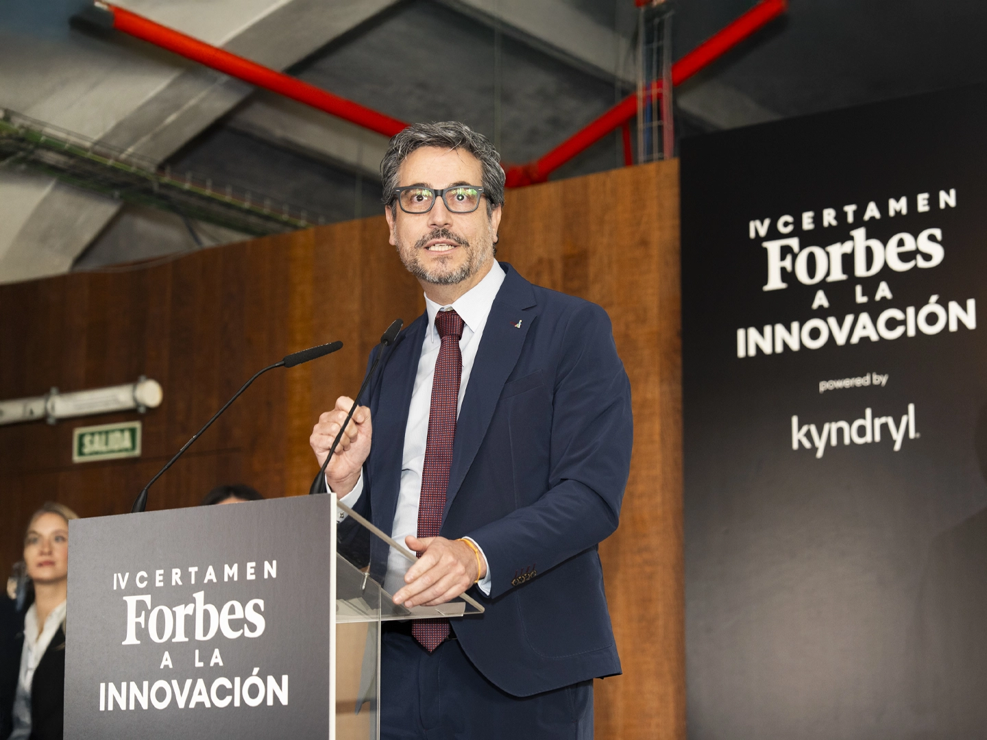 Hombre en podio con letras que ponen: "Certamen FORBES a la innovación" hablando con micrófono , vestido con traje azul marino y corbata