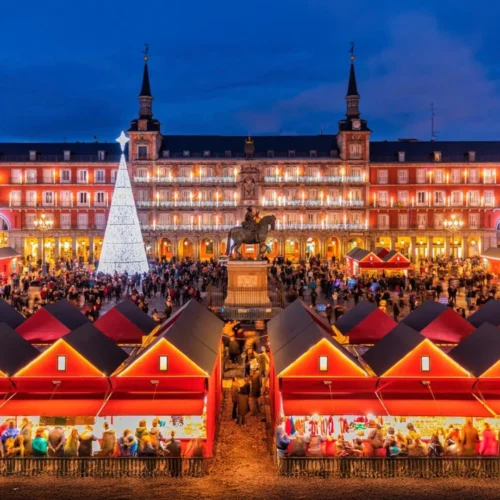 Mercado navideño iluminado en la Plaza Mayor de Madrid, con puestos rojos, un gran árbol de Navidad blanco y edificios históricos al fondo