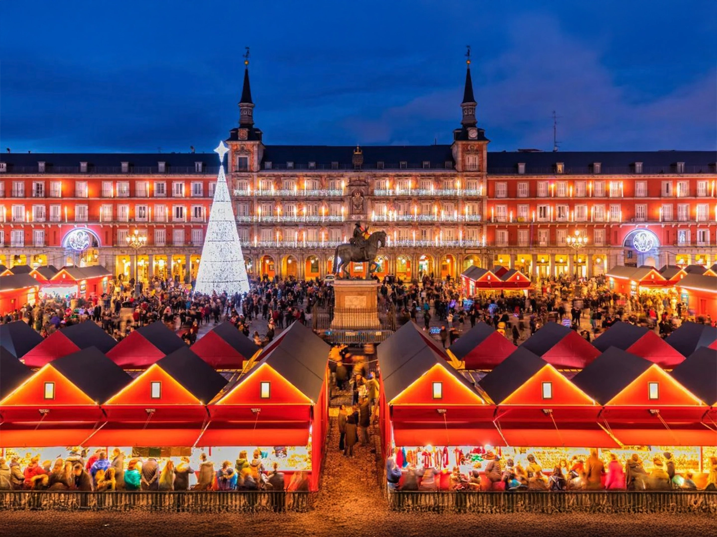Mercado navideño iluminado en la Plaza Mayor de Madrid, con puestos rojos, un gran árbol de Navidad blanco y edificios históricos al fondo