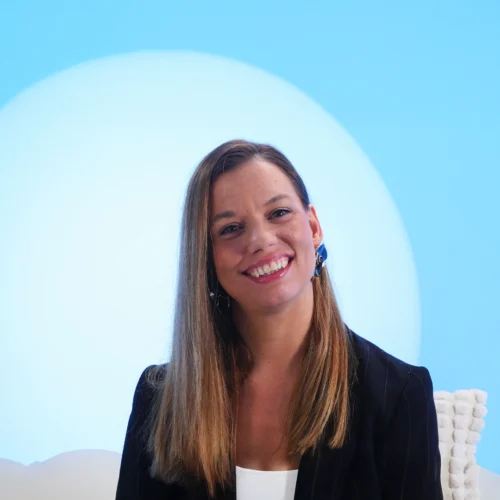 Mujer con cabello largo y liso, pendientes grandes y chaqueta de rayas oscuras, sonríe frente a un fondo azul claro con un círculo blanco detrás. La composición transmite cercanía, elegancia y profesionalismo, ideal para entrevistas, presentaciones o comunicación institucional.