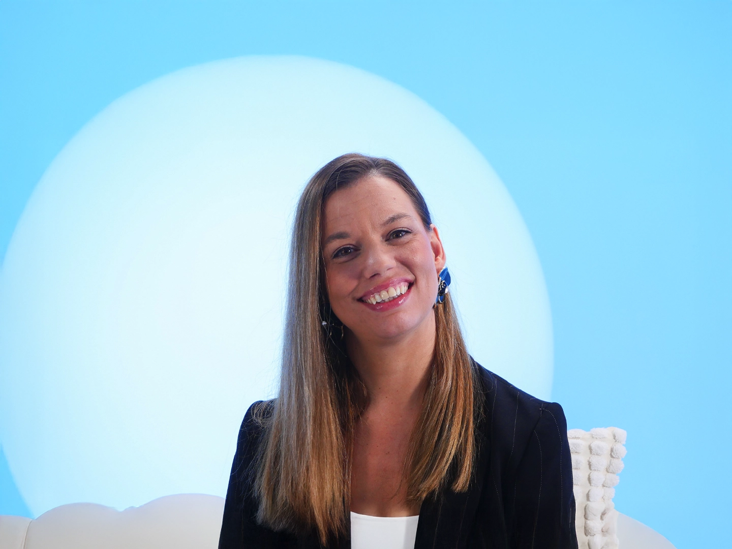 Mujer con cabello largo y liso, pendientes grandes y chaqueta de rayas oscuras, sonríe frente a un fondo azul claro con un círculo blanco detrás. La composición transmite cercanía, elegancia y profesionalismo, ideal para entrevistas, presentaciones o comunicación institucional.