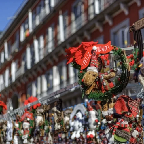 Puesto navideño al aire libre decorado con coronas, peluches y figuras festivas como renos y muñecos de nieve. Dos carteles con el mensaje “¡FELIZ NAVIDAD!” destacan entre los adornos. Al fondo, un edificio de ladrillo rojo con balcones y ventanas blancas refuerza el ambiente alegre y tradicional de las celebraciones navideñas en ciudad europea.
