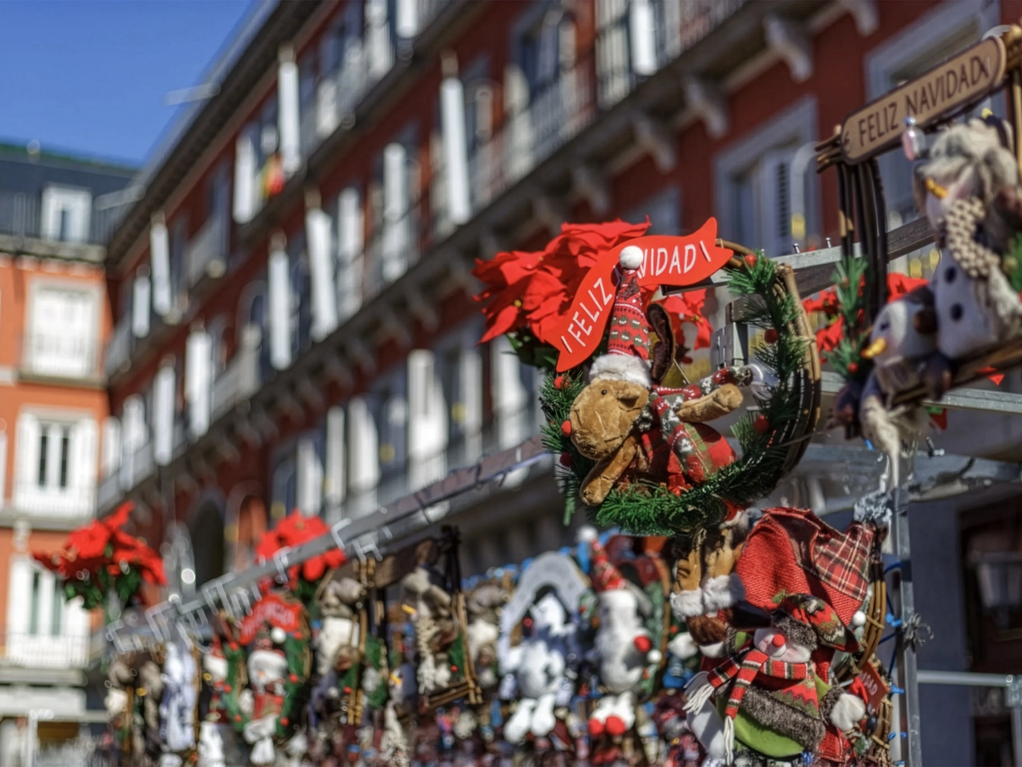 Puesto navideño al aire libre decorado con coronas, peluches y figuras festivas como renos y muñecos de nieve. Dos carteles con el mensaje “¡FELIZ NAVIDAD!” destacan entre los adornos. Al fondo, un edificio de ladrillo rojo con balcones y ventanas blancas refuerza el ambiente alegre y tradicional de las celebraciones navideñas en ciudad europea.