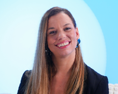 Retrato de una mujer sonriente con cabello largo y liso, pendientes azules y vestimenta oscura, sobre un fondo azul degradado con un círculo luminoso detrás. La composición transmite cercanía, elegancia y profesionalismo, ideal para uso corporativo o promocional.