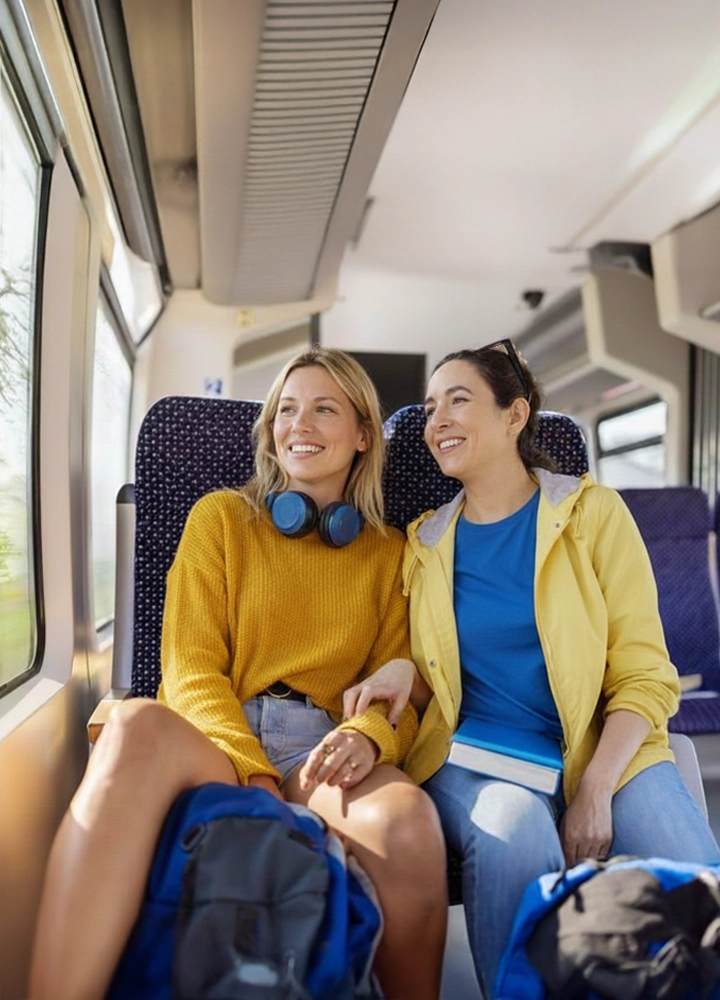 Dos mujeres sentadas en un tren, una con suéter amarillo y auriculares alrededor del cuello, la otra con camiseta azul y chaqueta amarilla, mochilas en el suelo.