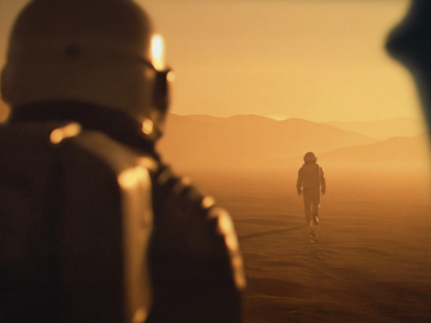 Primer plano del hombro de un astronauta en desenfoque, observando a otro astronauta que camina por un vasto paisaje desértico bajo una luz cálida y brumosa.