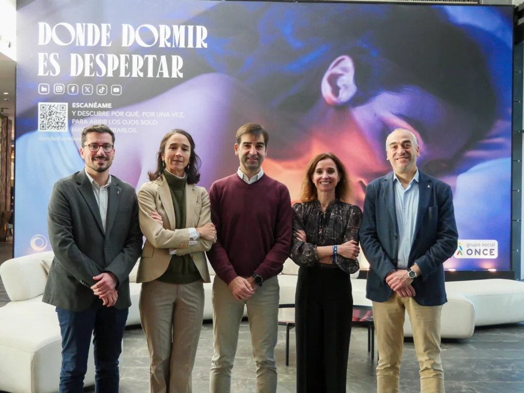 Cinco personas posando frente a un gran cartel promocional con el mensaje “Donde dormir es despertar” en un espacio interior moderno