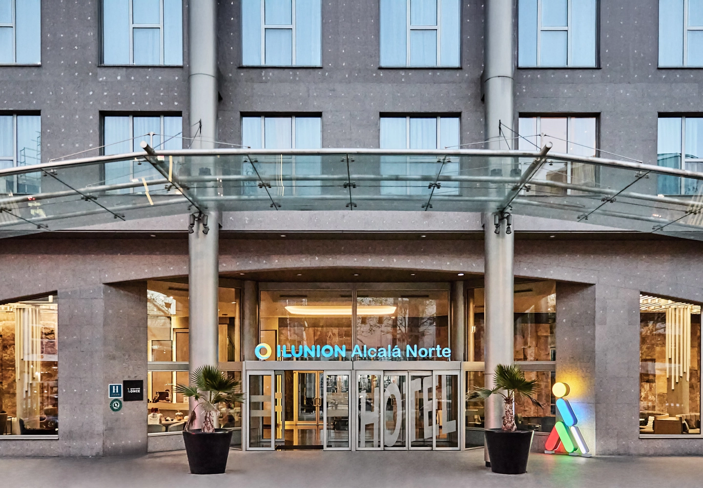 Entrada moderna de hotel con marquesina acristalada, plantas decorativas y luz natural que invita a una experiencia cómoda y accesible.