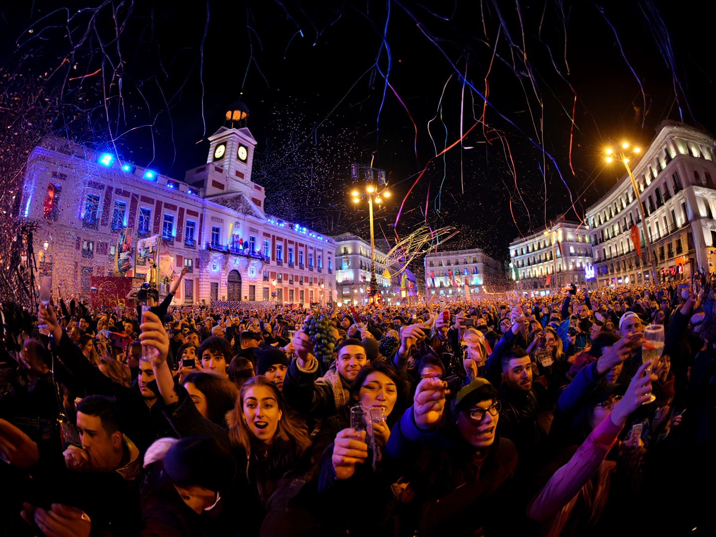 Multitud reunida frente a la Real Casa de Correos en la Puerta del Sol, Madrid, celebrando la Nochevieja. El reloj iluminado marca la cuenta atrás hacia la medianoche, mientras los asistentes sostienen uvas y copas, siguiendo la tradición española de tomar doce uvas al ritmo de las campanadas para atraer la buena suerte. La escena está llena de confeti, serpentinas y luces festivas, transmitiendo alegría colectiva y el valor cultural de esta celebración emblemática.