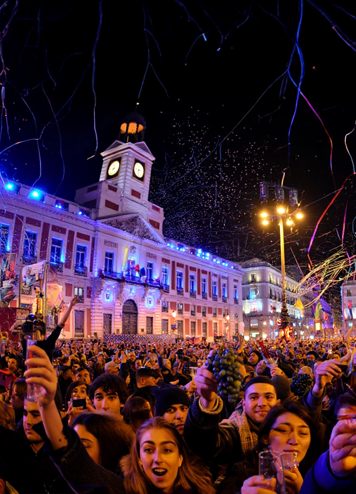 Multitud reunida frente a la Real Casa de Correos en la Puerta del Sol, Madrid, celebrando la Nochevieja. El reloj iluminado marca la cuenta atrás hacia la medianoche, mientras los asistentes sostienen uvas y copas, siguiendo la tradición española de tomar doce uvas al ritmo de las campanadas para atraer la buena suerte. La escena está llena de confeti, serpentinas y luces festivas, transmitiendo alegría colectiva y el valor cultural de esta celebración emblemática.