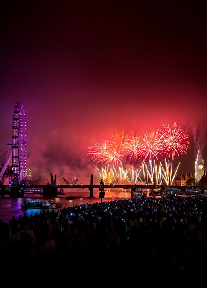 Espectáculo de fuegos artificiales sobre el río Támesis en Londres, con el London Eye iluminado en tonos púrpura a la izquierda y el Big Ben visible a la derecha. Una multitud se reúne en la ribera, muchos capturando el momento con sus móviles. Los fuegos artificiales en rojo, amarillo y blanco iluminan el cielo nocturno, creando una atmósfera festiva y vibrante. La imagen transmite celebración, emoción colectiva y el poder simbólico de los íconos urbanos.