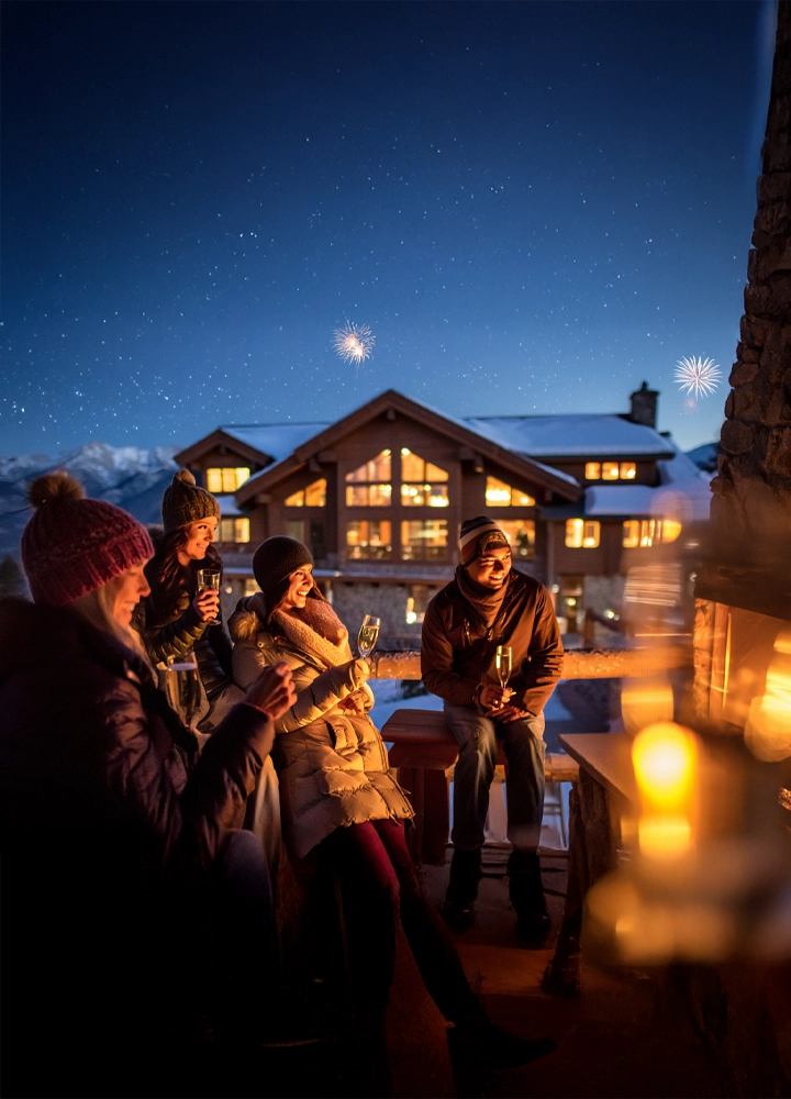 Escena nocturna en un entorno invernal, con cuatro personas abrigadas reunidas alrededor de una chimenea exterior, disfrutando de bebidas y compañía. Al fondo, una cabaña de madera iluminada y techos cubiertos de nieve se integra en un paisaje montañoso bajo un cielo estrellado. Los fuegos artificiales en el cielo sugieren una celebración.
