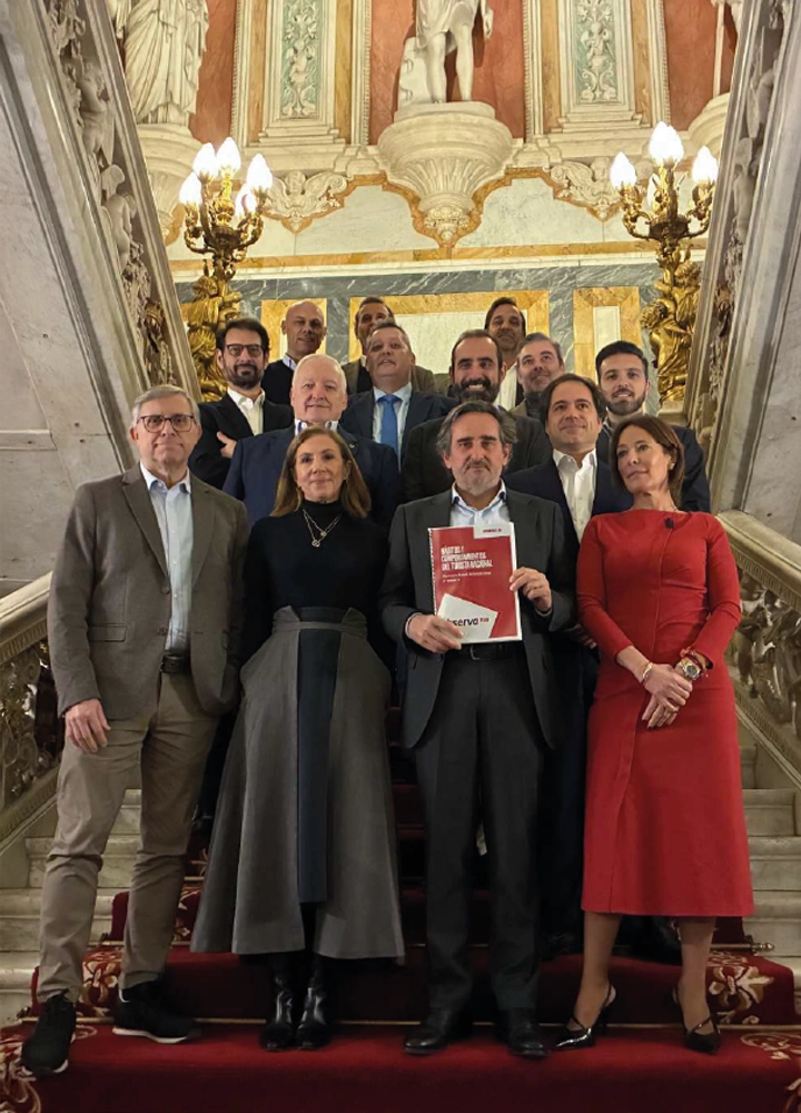 Grupo de quince personas posando en una escalera monumental dentro de un edificio de gran riqueza arquitectónica. El entorno, con barandillas de mármol, candelabros dorados y detalles escultóricos, transmite solemnidad y elegancia. En el centro, una persona sostiene una carpeta roja y blanca con texto, lo que sugiere un acto oficial vinculado a comunicación institucional o patrimonio cultural.