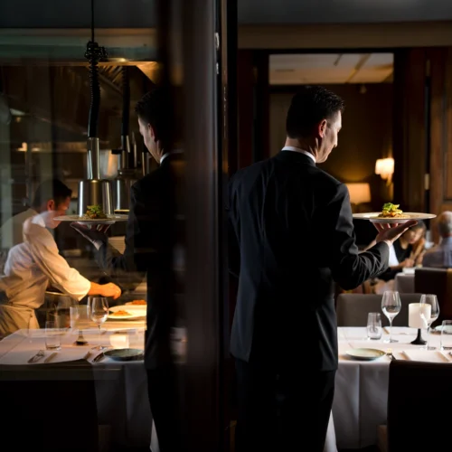 Escena de restaurante de alta cocina con un camarero vestido de traje oscuro sirviendo un plato gourmet. Al fondo, la cocina visible tras una mampara de cristal muestra a un chef trabajando bajo lámparas de calor. El comedor está elegantemente dispuesto con manteles blancos, copas de vino y velas, mientras los comensales conversan en un ambiente refinado. La imagen refleja la armonía entre el dinamismo del servicio y la experiencia serena del cliente.