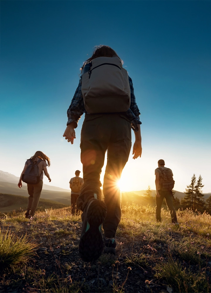 Grupo de cuatro senderistas caminando por un paisaje de hierba durante el amanecer o atardecer. La perspectiva desde atrás destaca la mochila y las botas de uno de ellos. La luz cálida del sol bajo proyecta sombras alargadas sobre el terreno, mientras se aprecian árboles y picos montañosos en la distancia. La imagen transmite aventura, conexión con la naturaleza y disfrute activo del entorno, ideal para comunicar valores de bienestar, sostenibilidad y turismo responsable.