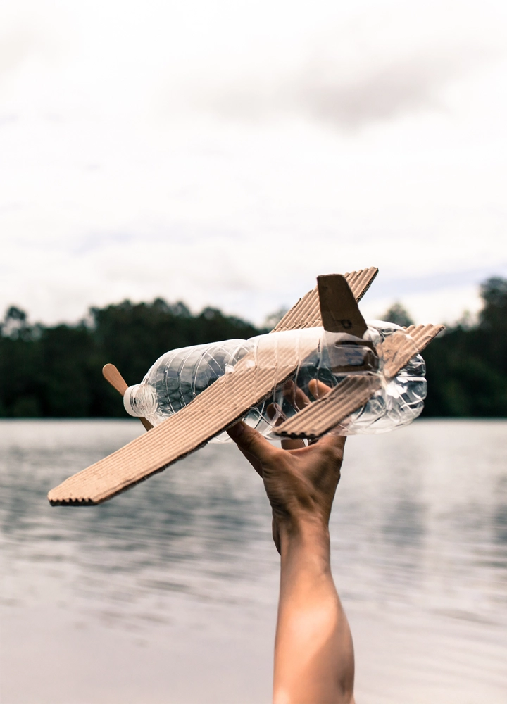 Mano sosteniendo un avión de juguete hecho a mano con materiales reciclados: el cuerpo es una botella de plástico transparente, mientras que las alas, la cola y la hélice están construidas con cartón y palitos de madera. Al fondo, un paisaje natural con agua tranquila, árboles y cielo nublado. La imagen transmite creatividad, sostenibilidad y juego consciente, ideal para comunicar valores educativos, medioambientales o iniciativas de reutilización.