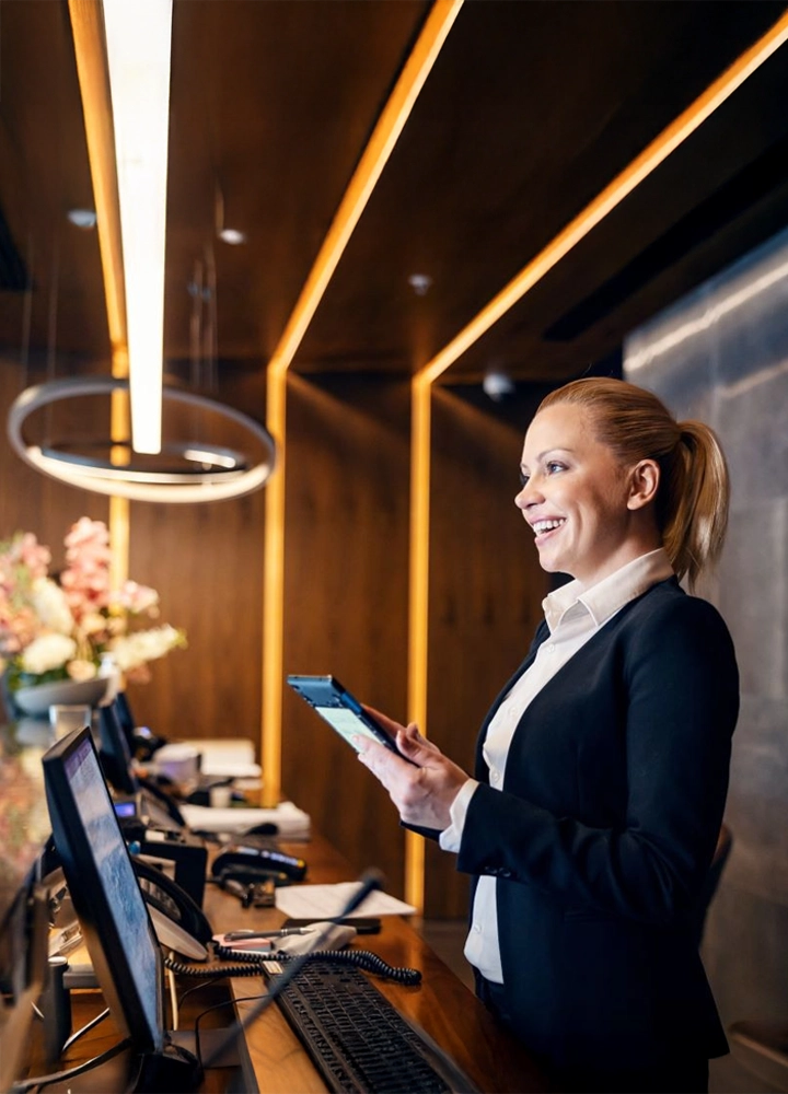 Mujer en mostrador de recepción, sosteniendo una tablet en un entorno elegante con iluminación cálida y decoración floral