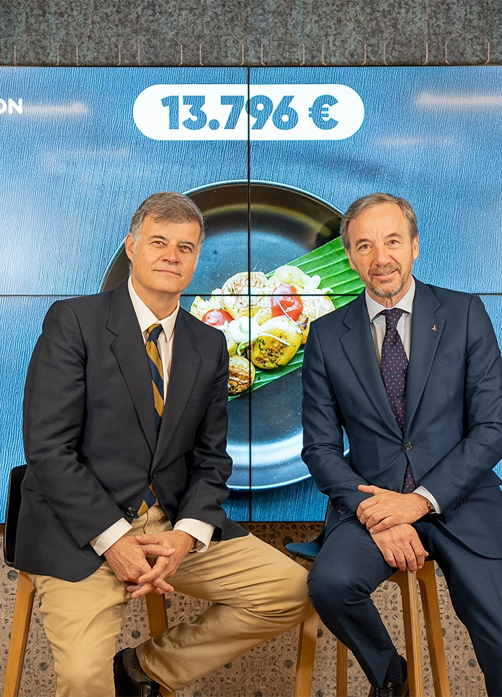 Plano medio de dos hombres sentados en taburetes frente a una pantalla digital que muestra una imagen de comida y la cifra 