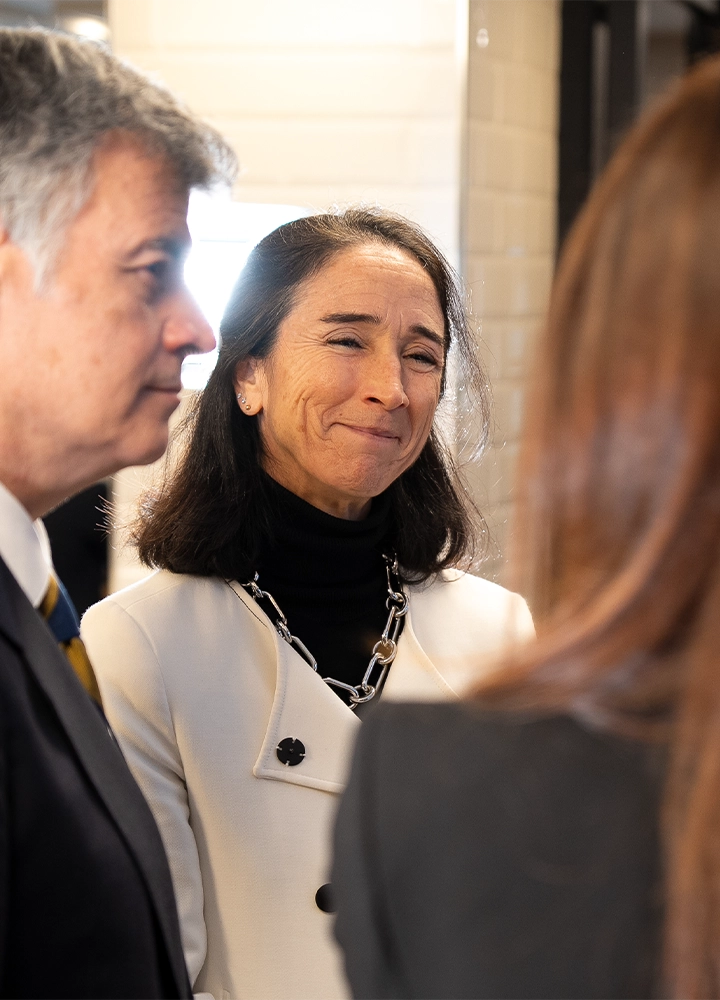 Primer plano de una mujer de cabello oscuro con una expresión de emoción o gratitud mientras conversa con otras personas, de quienes solo se ven partes del rostro o el hombro. Viste una chaqueta blanca sobre un jersey de cuello alto negro y un collar de cadena plateada. El fondo es una pared de azulejos claros.