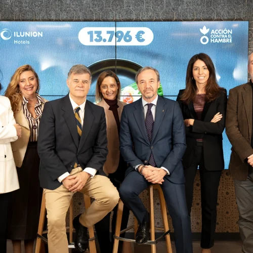 Un grupo de siete personas posa frente a una gran pantalla digital. Dos hombres están sentados en taburetes de madera en el centro, flanqueados por cinco personas de pie. La pantalla muestra los logotipos de 