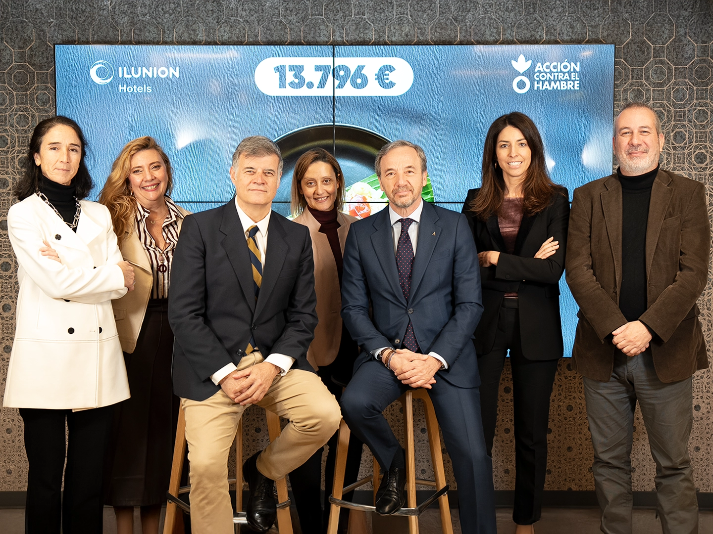 Un grupo de siete personas posa frente a una gran pantalla digital. Dos hombres están sentados en taburetes de madera en el centro, flanqueados por cinco personas de pie. La pantalla muestra los logotipos de "ILUNION Hotels" y "ACCIÓN CONTRA EL HAMBRE", junto con la cifra "13.796 €" destacada en una burbuja blanca. Todos visten atuendos de negocios o casual elegante y sonríen a la cámara. La pared del fondo tiene un patrón geométrico oscuro.