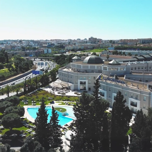 Vista aérea del Ilunion Hotels en un entorno urbano, destacando una piscina exterior de color azul brillante rodeada de jardines con palmeras y sombrillas. Al fondo se observa una autovía con tráfico y el horizonte de la ciudad bajo un cielo despejado.