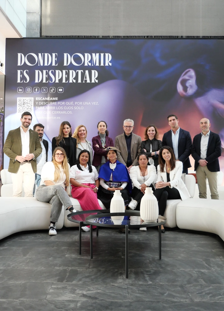 Fotografía grupal de un equipo de 14 personas posando delante de un gran mural publicitario. El mural muestra el rostro de una persona durmiendo y el texto en letras grandes: 'DONDE DORMIR ES DESPERTAR', seguido de iconos de redes sociales y un código QR con el mensaje 'Escanéame y descubre por qué, por una vez, para abrir los ojos solo hay que cerrarlos'. El grupo, compuesto por hombres y mujeres con vestimenta profesional y una mujer con traje tradicional andino, está distribuido de pie y sentado en un sofá blanco curvo en un entorno moderno.