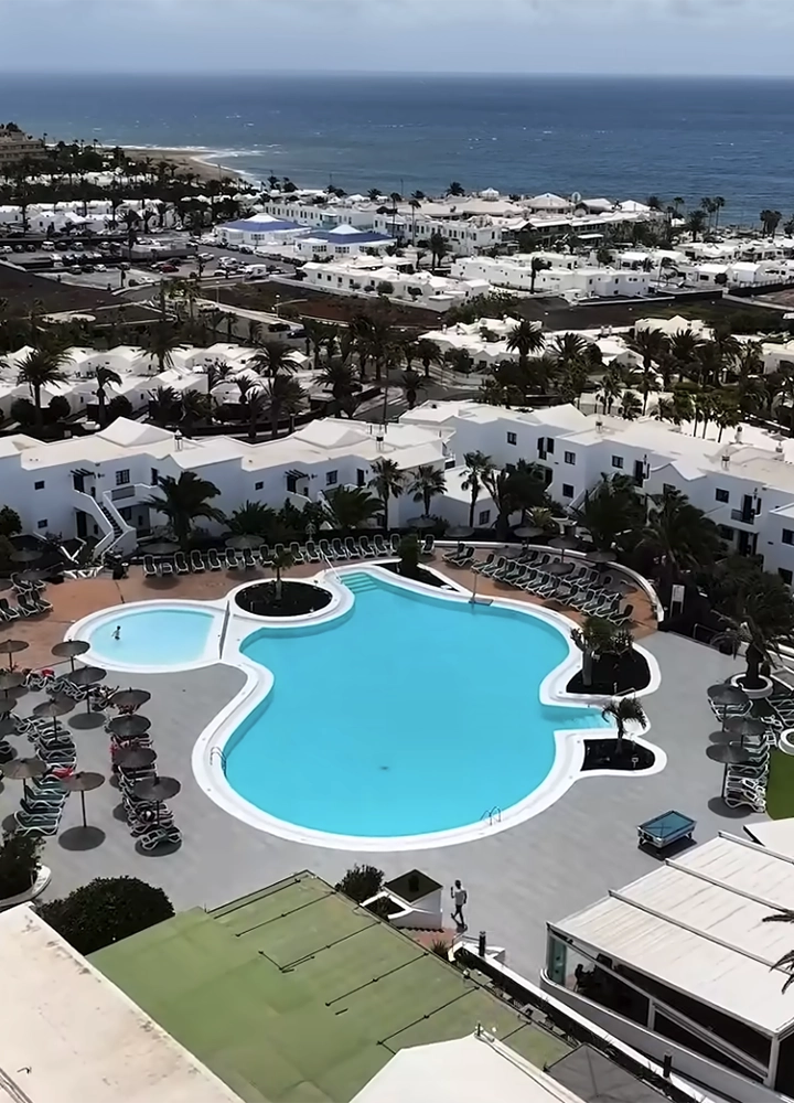 Una fotografía aérea de un complejo turístico costero con una gran piscina central de color azul turquesa y formas curvas. El resort está compuesto por edificios de arquitectura blanca y baja, rodeados de palmeras y zonas de tumbonas. Al fondo, se aprecia una playa.