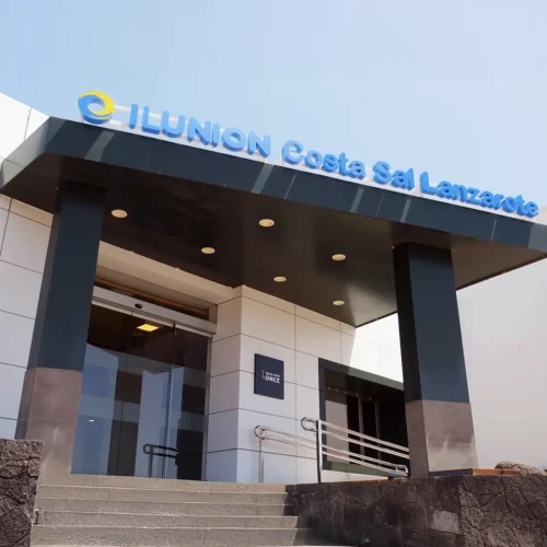 Fotografía de la entrada principal del hotel ILUNION Costa Sal Lanzarote. Se muestra una estructura moderna con una fachada de paneles blancos y una gran marquesina negra sobre la puerta de cristal automática. En la parte superior de la marquesina, destaca el rótulo con letras azules iluminadas 