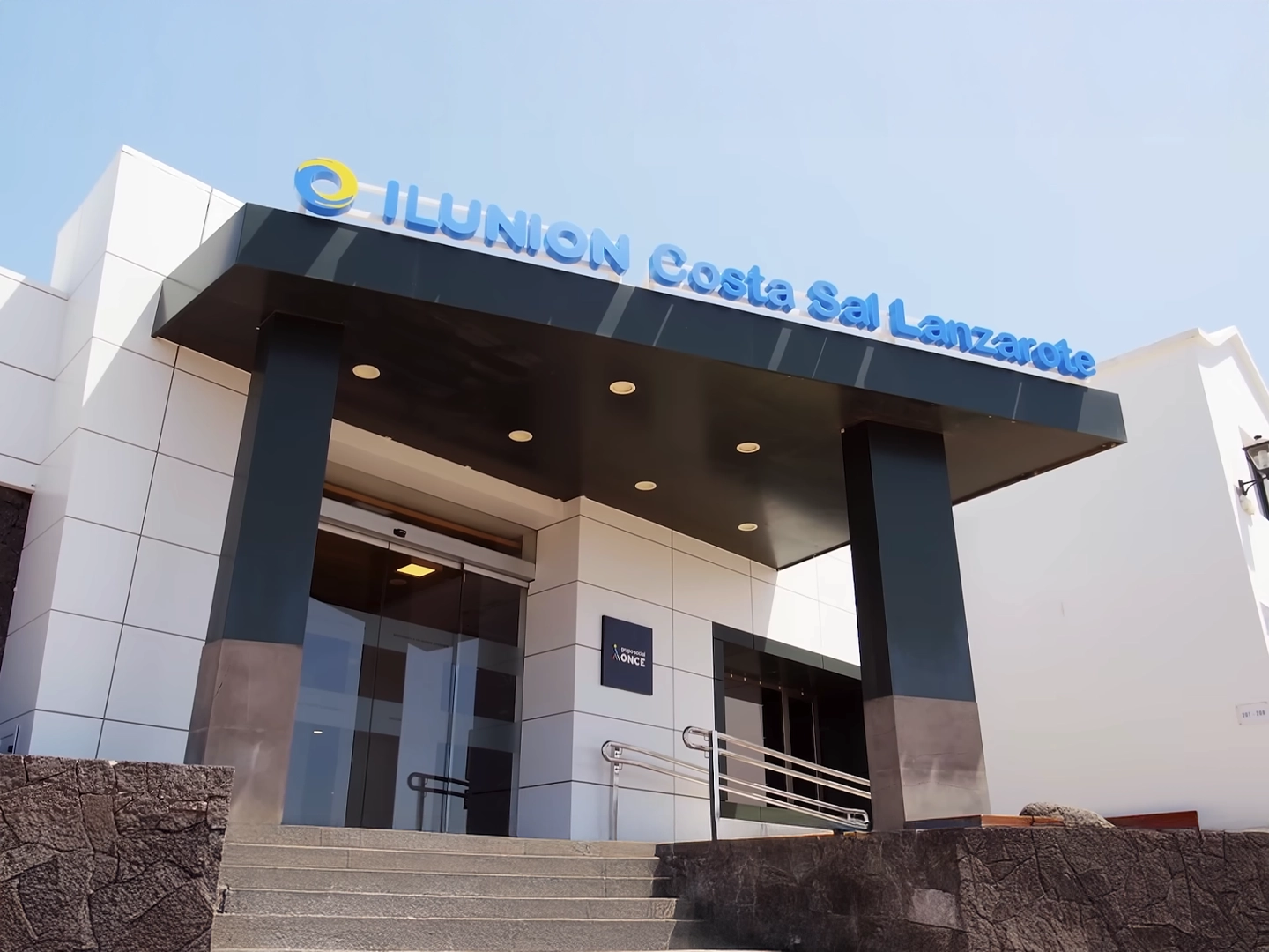 Fotografía de la entrada principal del hotel ILUNION Costa Sal Lanzarote. Se muestra una estructura moderna con una fachada de paneles blancos y una gran marquesina negra sobre la puerta de cristal automática. En la parte superior de la marquesina, destaca el rótulo con letras azules iluminadas "ILUNION Costa Sal Lanzarote" junto a un logotipo circular amarillo y azul. Unas escaleras de piedra gris y una rampa con barandilla metálica conducen al acceso principal bajo un cielo despejado.