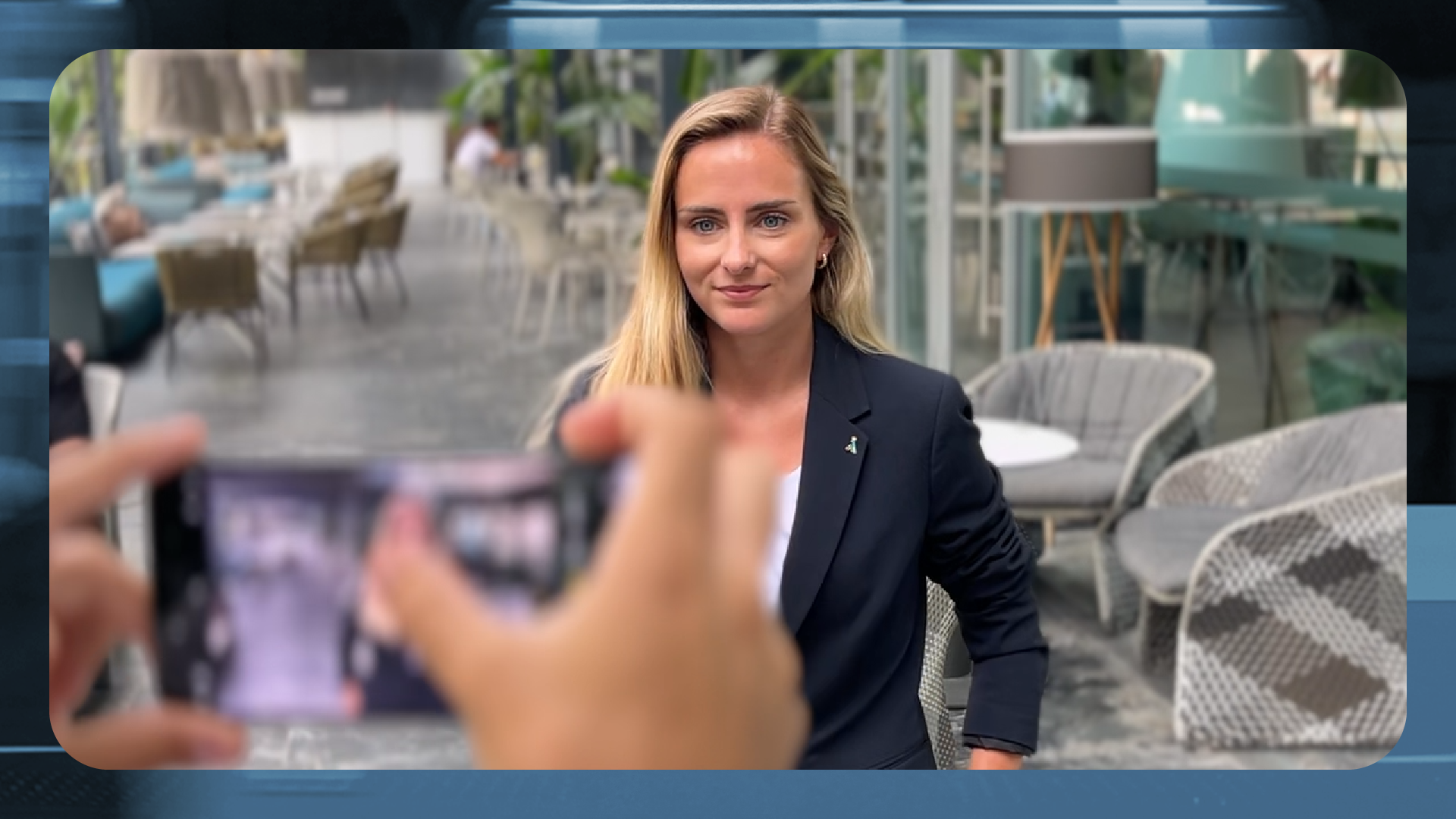 Fotografía de una mujer rubia con chaqueta de traje azul oscuro sentada en una terraza moderna de un hotel. Mira directamente a la cámara con una sonrisa leve y profesional. En primer plano, unas manos desenfocadas sostienen un smartphone que la está grabando o fotografiando. El fondo muestra un entorno amplio con mesas, sillas de diseño y plantas, creando una atmósfera de entrevista o reportaje corporativo.