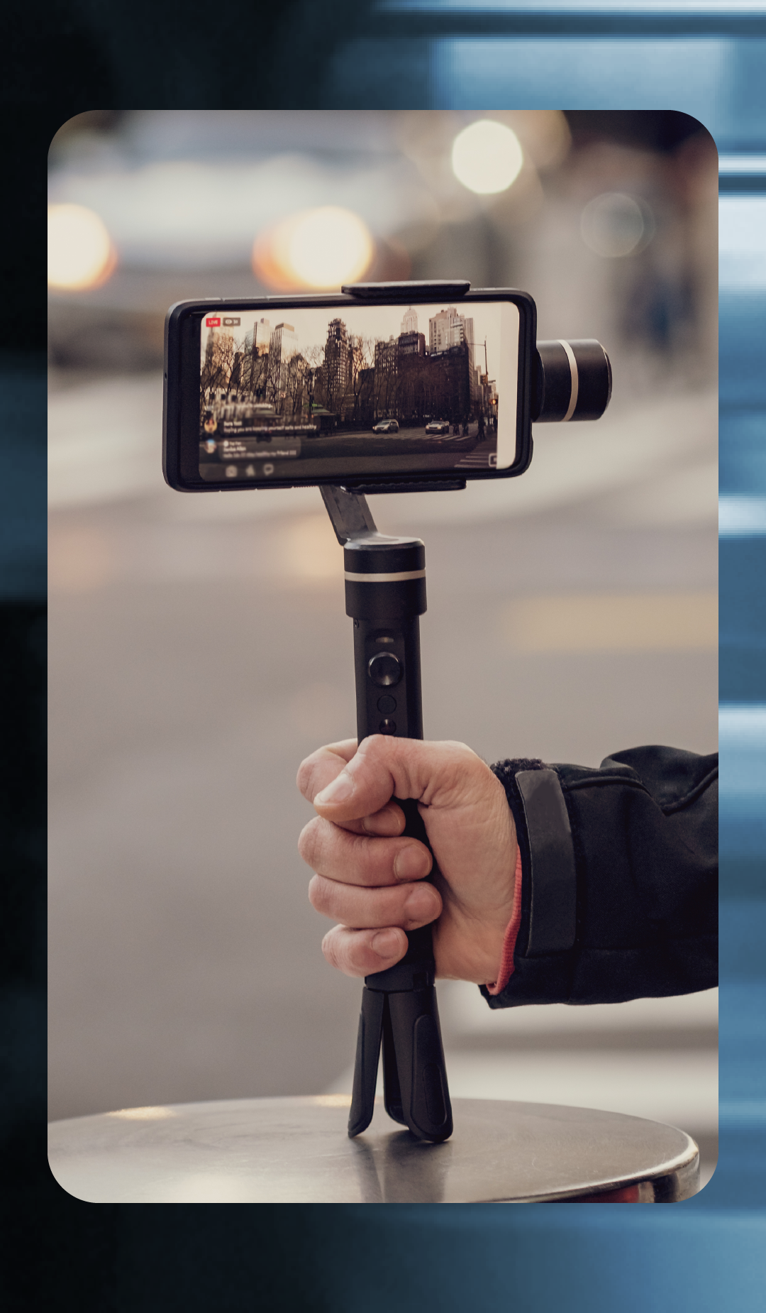 Primer plano de una mano sosteniendo un estabilizador de mano (gimbal) para smartphone apoyado sobre una superficie metálica circular. En la pantalla del teléfono se observa una transmisión en vivo que muestra una calle con edificios altos de una gran ciudad y tráfico de vehículos. El fondo de la imagen real está desenfocado, sugiriendo un entorno urbano exterior con luces cálidas.