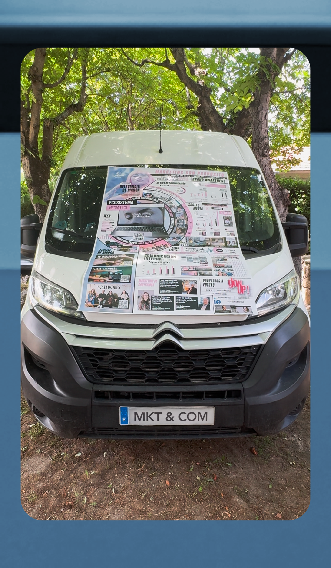 Fotografía frontal de una furgoneta blanca marca Citroën estacionada bajo unos árboles. Sobre el capó y el parabrisas hay desplegado un gran cartel publicitario o infografía de marketing con el título 'Marketing con Propósito'. El cartel incluye secciones como 'Ecosistema Mediático', 'Social Media', y 'Proyectos a Futuro', además de gráficos, fotos y un código QR. En el centro del diseño aparece un portátil con la frase 'Donde dormir es despertar' (que se traduce como 'where sleeping is an awakening'). La matrícula de la furgoneta ha sido personalizada con el texto 'MKT &amp; COM'.