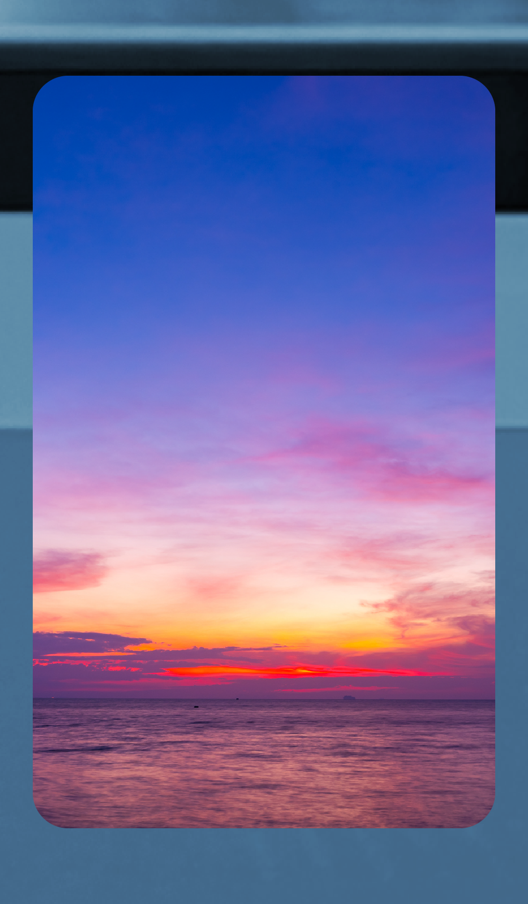 Fotografía vertical de una puesta de sol sobre un mar en calma. El cielo muestra un degradado vibrante que va desde un azul profundo en la parte superior, pasando por tonos violetas y rosas, hasta llegar a un naranja y rojo intenso en el horizonte. El agua refleja los colores cálidos del atardecer con suaves ondulaciones, creando una atmósfera serena y contemplativa.