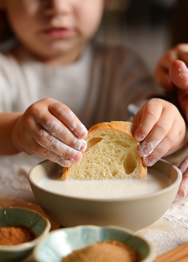 Primer plano de las manos de un niño pequeño, cubiertas de harina, mojando una rebanada de pan artesanal en un cuenco con leche. En la esquina inferior izquierda se ven dos pequeños recipientes con canela y azúcar moreno sobre una mesa enharinada. Al fondo, la figura desenfocada del niño sugiere una actividad de cocina en familia, con una iluminación suave y natural que crea una atmósfera acogedora.