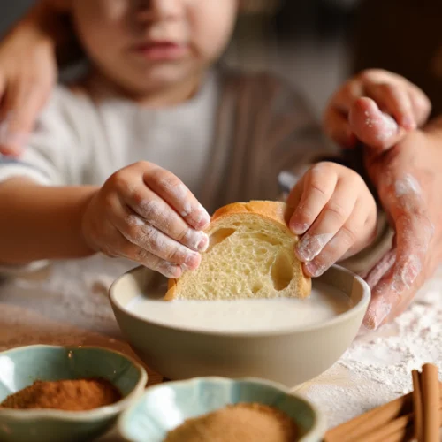 Primer plano de las manos de un niño pequeño, cubiertas de harina, mojando una rebanada de pan artesanal en un cuenco con leche. En la esquina inferior izquierda se ven dos pequeños recipientes con canela y azúcar moreno sobre una mesa enharinada. Al fondo, la figura desenfocada del niño sugiere una actividad de cocina en familia, con una iluminación suave y natural que crea una atmósfera acogedora.