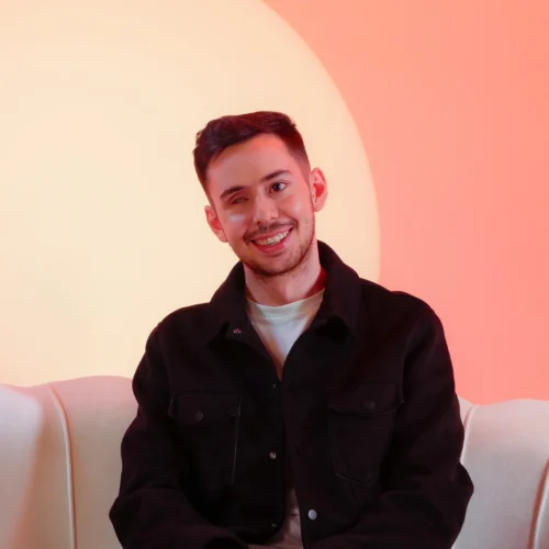 Retrato de un hombre joven sonriente sentado en un sofá blanco de estilo moderno con respaldo ondulado. Viste una chaqueta oscura sobre una camiseta clara. El fondo es minimalista con una iluminación en tonos cálidos anaranjados y amarillos que forman un círculo de luz detrás de él.